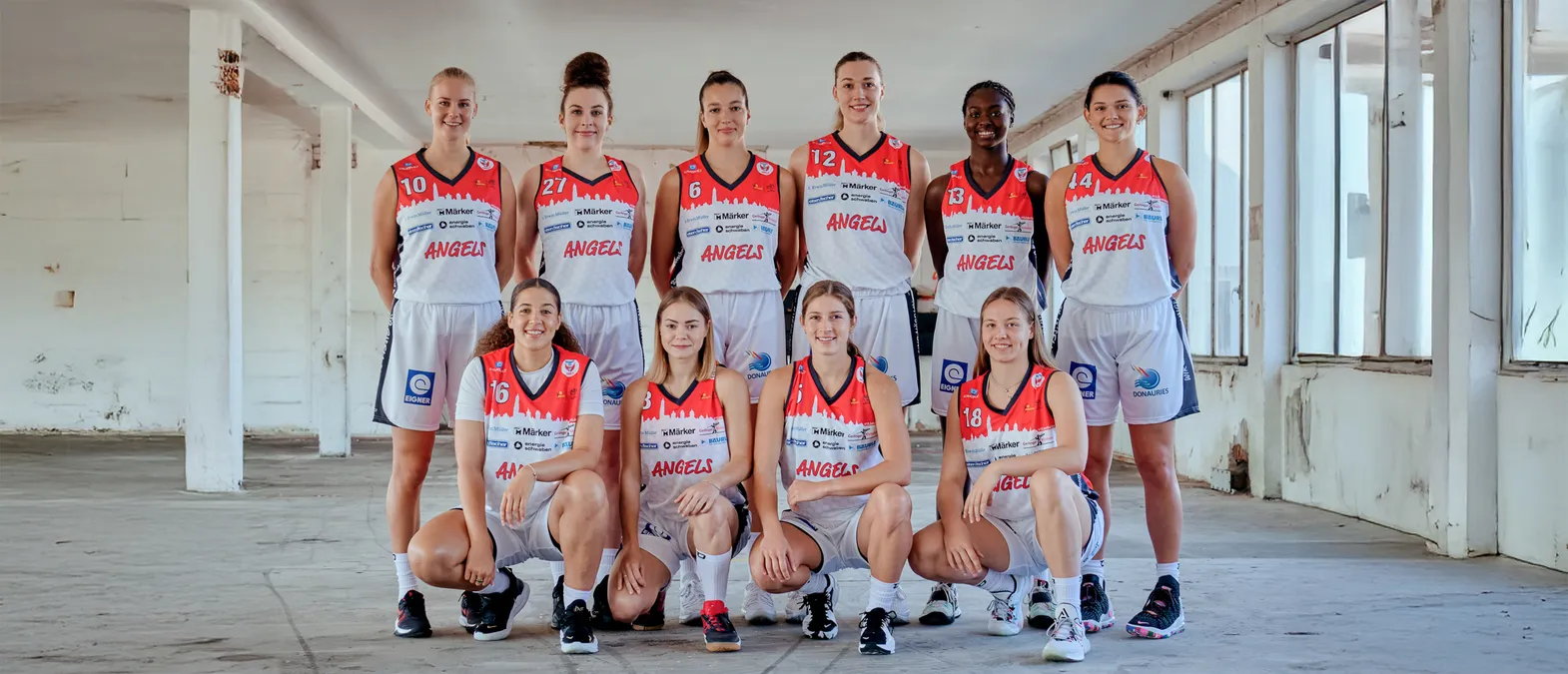 En gruppe med åtte kvinner i oransje og hvite basketballtrøyer med ordet 'ANGELS' på poserer i et lager.