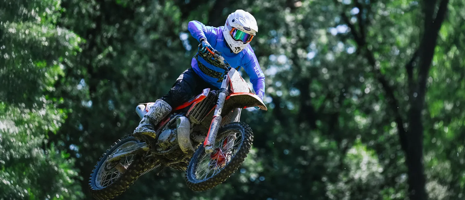 En motorsyklist i blå, langarmet racingsett og hvit hjelm hopper på motorsykkel gjennom en skog.