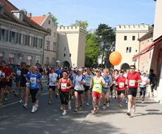 En gruppe løpere i sportstøy løper nedover en bygate, hvor noen holder en oransje ballong.