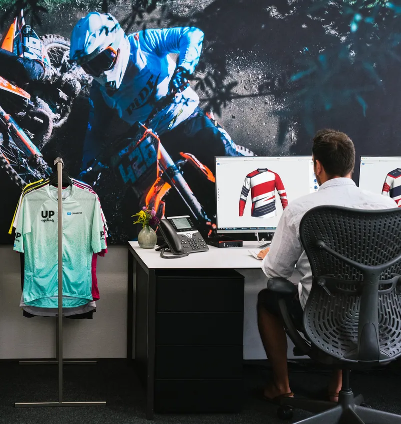 En mann sitter ved et skrivebord og designer et tilpasset sportskjole på en datamaskin, mens en stor bilde av en motorsyklist og en klærhenger med ulike sportsklær er synlig i bakgrunnen.