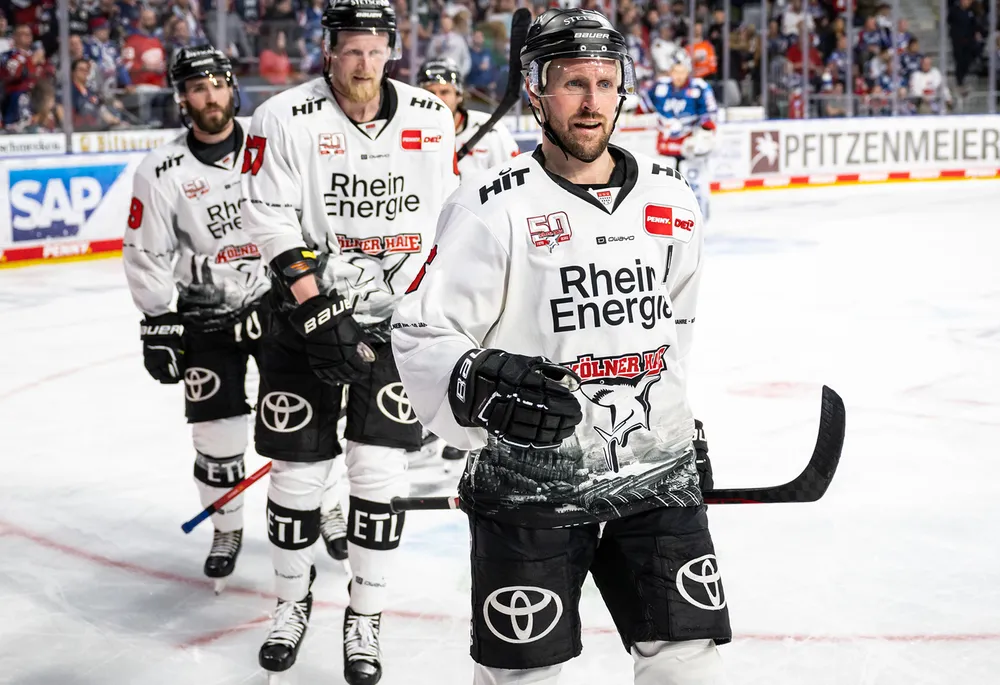 Ishockeyspillere i hvite trøyer med svarte bukser som har et laglogo, stående på isen.