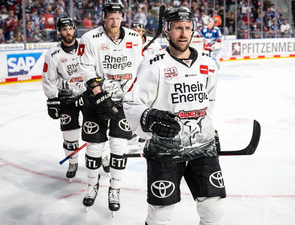 Tre ishockeyspillere i hvite trøyer med svarte logoer og grafiske design står på isen.