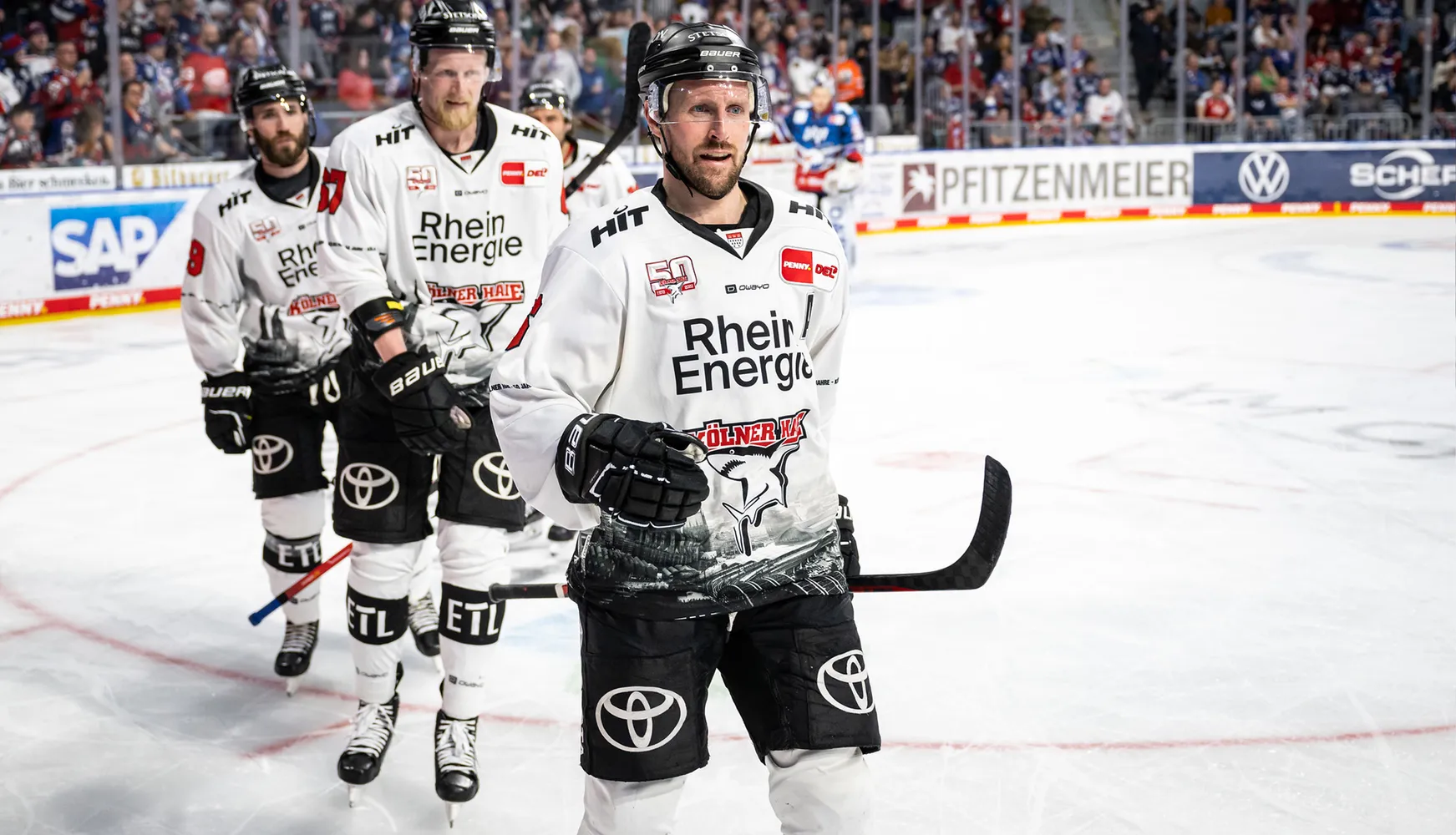 Tre ishockeyspillere i hvite trøyer med forskjellige nummer står på isen under en kamp.