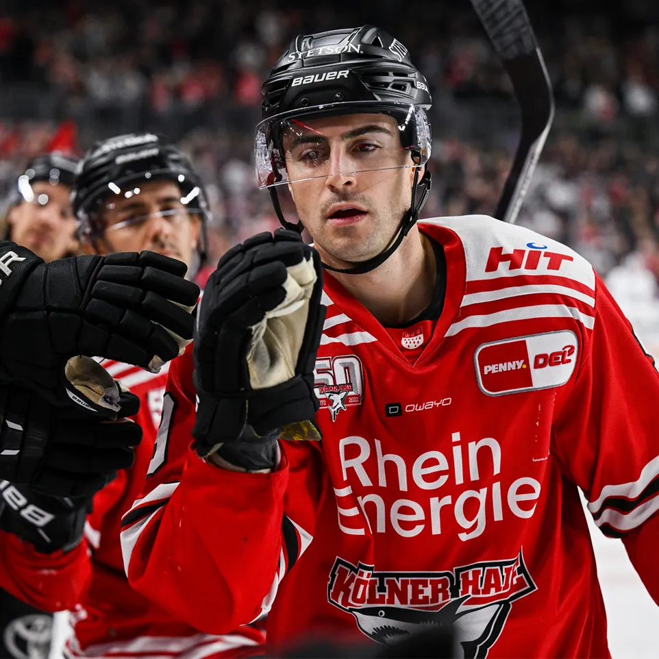 En hockeyspiller i en rød trøye med hvite striper og logoer, som hever hånden i feiring.