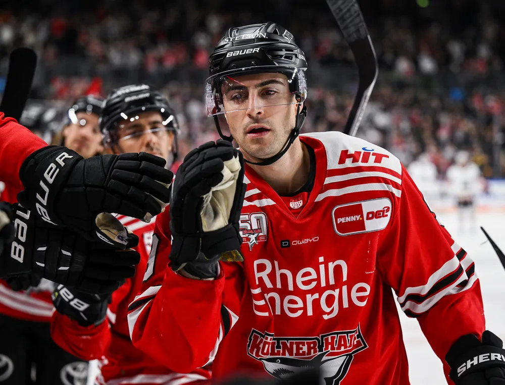 En hockeyspiller har på seg en rød trøye med hvite og svarte detaljer mens han interagerer med en lagkamerat.