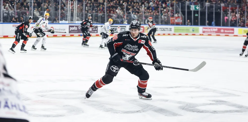 En ishockeyspiller i en svart trøye med røde og hvite detaljer som går på isen med en kølle, mens publikum jubler i bakgrunnen.