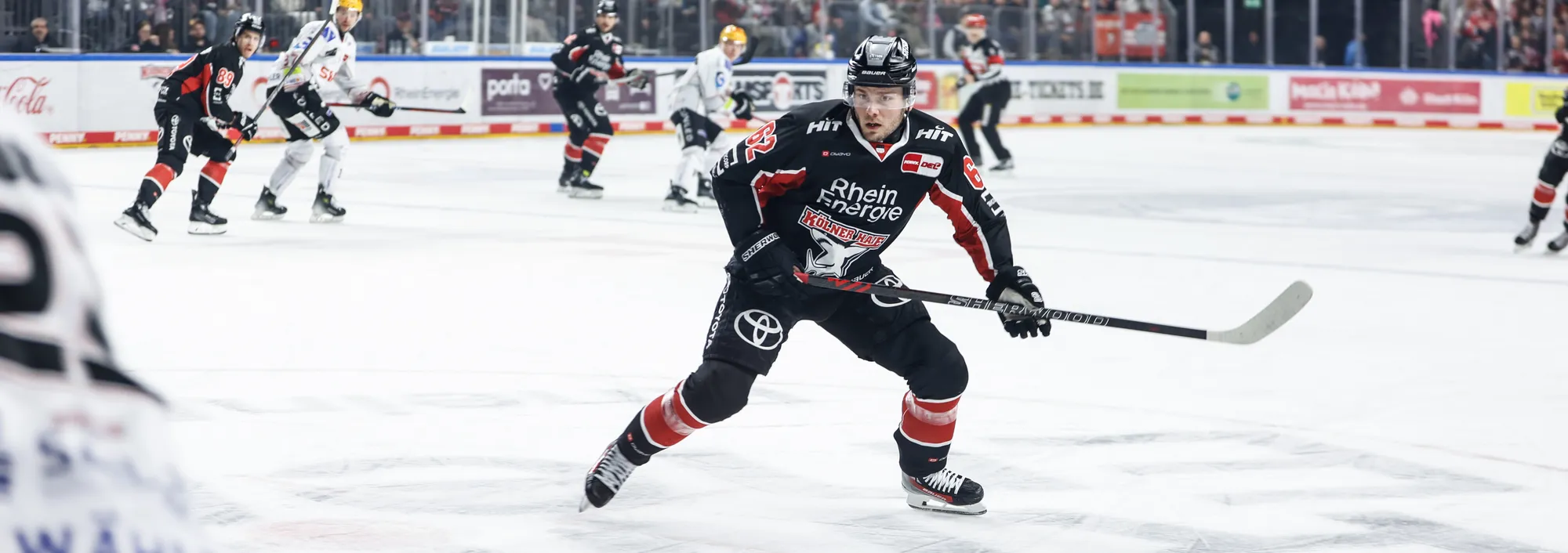 En ishockeyspiller i en svart og rød trøye som står på isen med en hockeykølle.