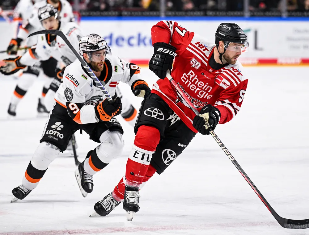 En ishockeyspiller i rød trøye og svarte bukser går på isen mens han blir forfulgt av en annen spiller.