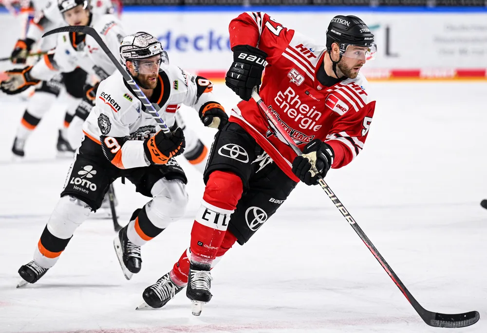En hockeyspiller i en rød trøye står på skøyter med pucken på isen, mens en annen spiller i en hvit trøye følger tett bak.