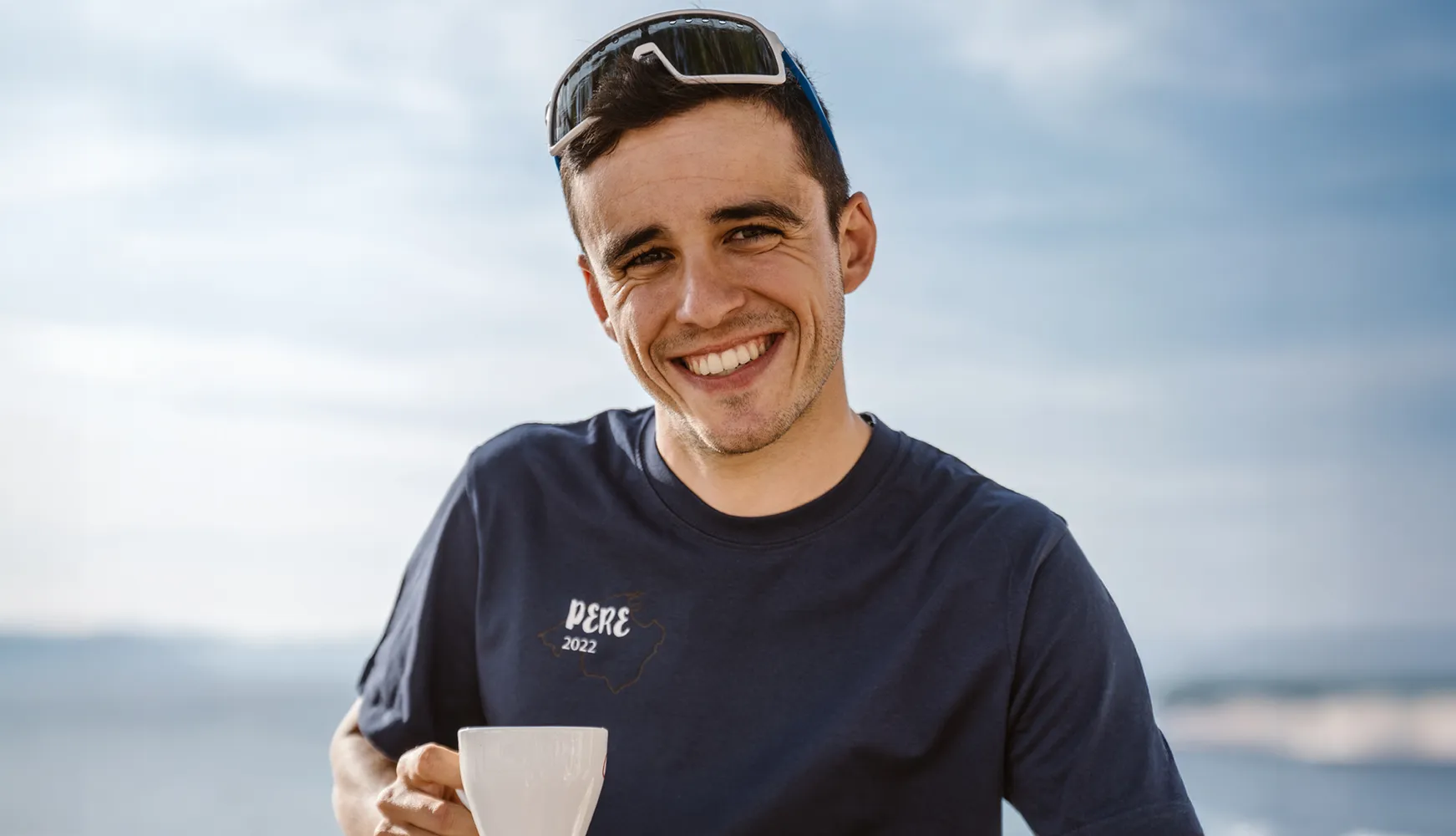En ung mann smiler i en blå T-skjorte med teksten 'PERE 2022' mens han holder en kopp, mot en klar himmel.