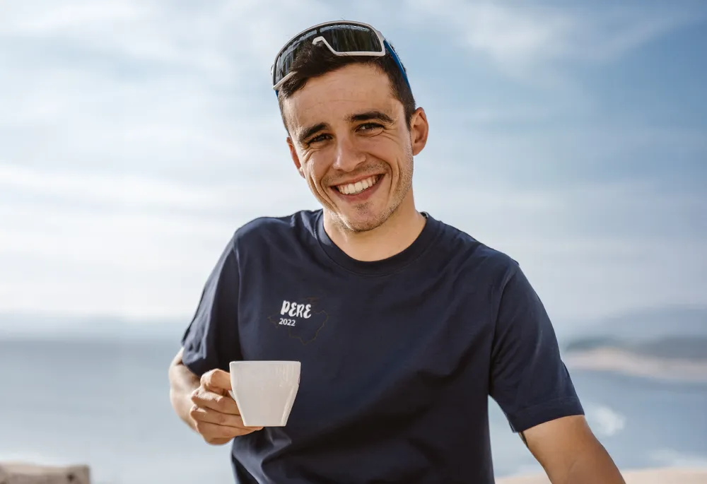 En smilende mann i en blå T-skjorte holder en kopp i hånden, med et vakkert kystlandskap i bakgrunnen.