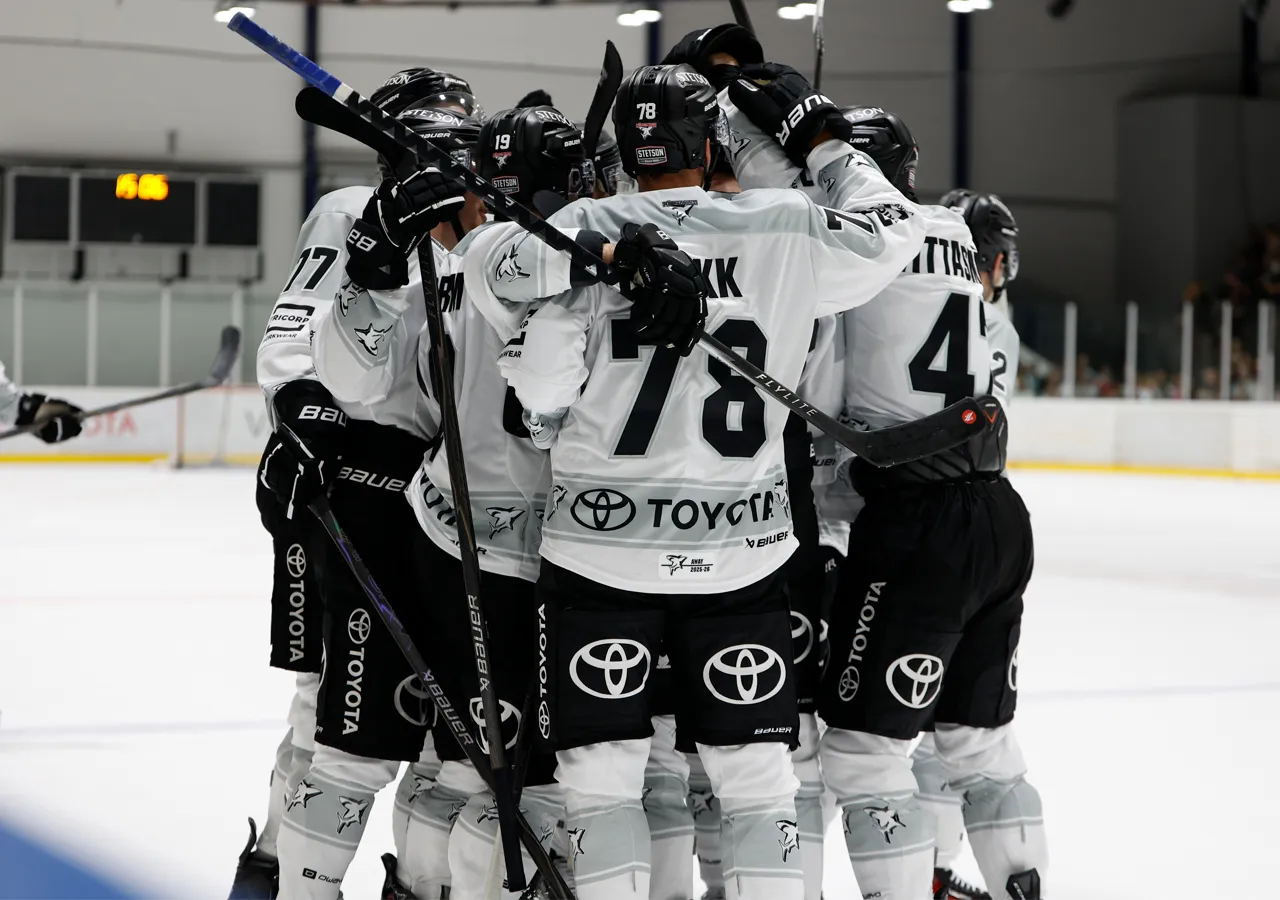 Hockeyspillere i hvite trøyer med svarte numre og sponsorlogoer som klemmer hverandre på isen.