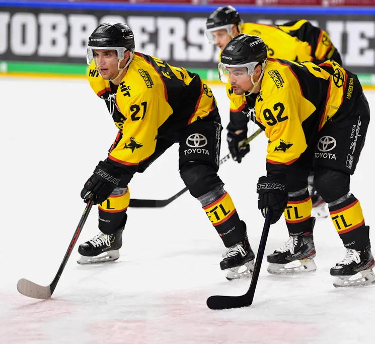 Hockeyspillere i gule og svarte drakter med nummer 21 og 92 på isen, som holder sine staver.