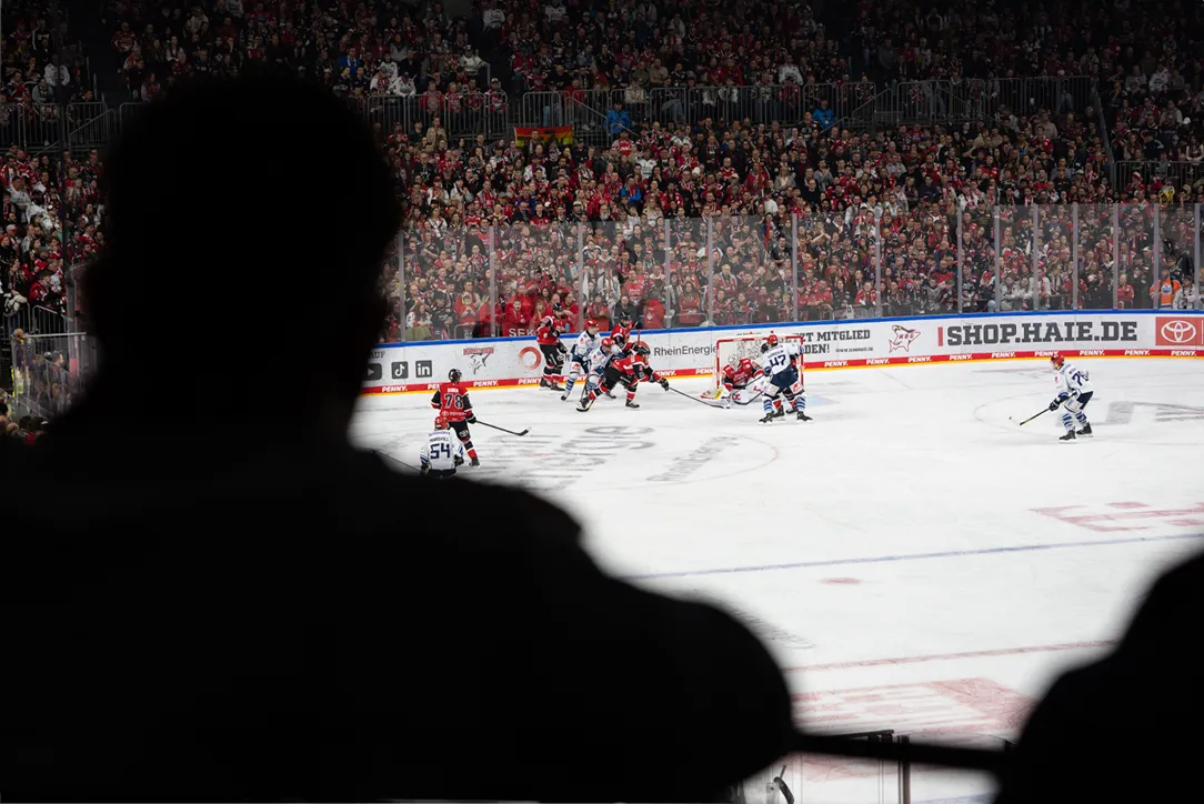 Fra et tilskuers synspunkt viser bildet en ishockeykamp med spillere i røde og blå drakter foran en stor folkemengde.