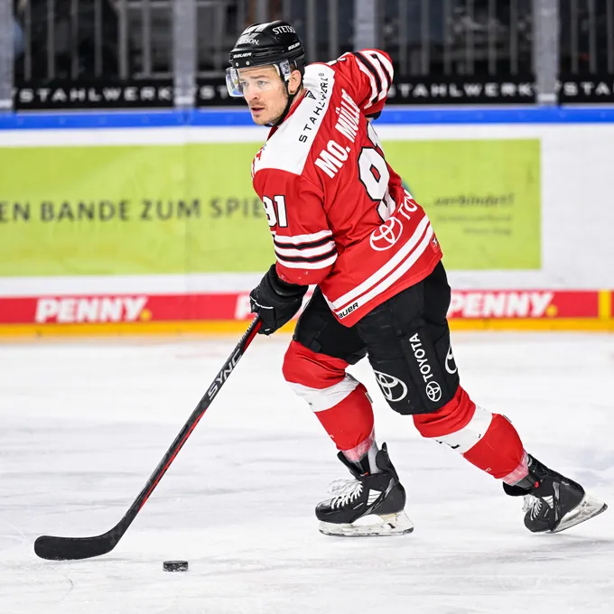 En ishockeyspiller i en rød og svart trøye med nummer 91 og navnet Mc. Miller på ryggen, stående på isen og kontrollerer en puck med sin stok.