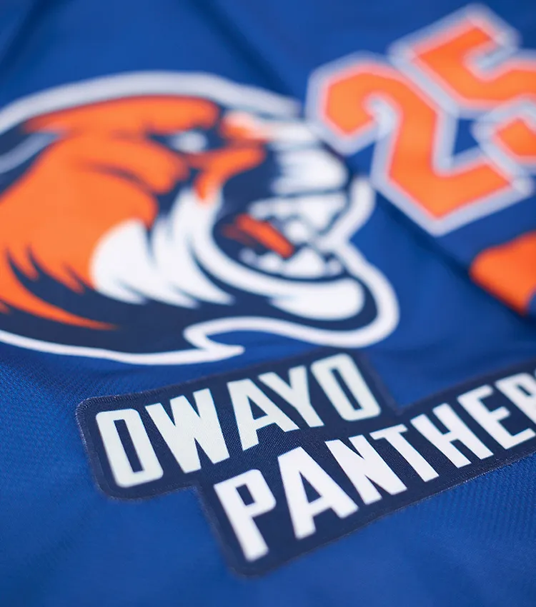 Nærbeskrivelse av en blå sportskjorte med et oransje panterlogo og teksten 'OWAYO PANTHERS' i hvite bokstaver på en mørk plakett.