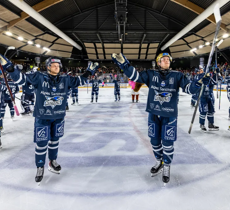 Hockeyspillere i blå trøyer med sponsorlogoer feirer på isen, mens publikum er synlig i bakgrunnen.