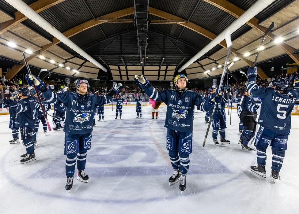 Ishockeyspillere i blå trøyer står på isen og feirer med hevede køller foran et jubelende publikum.