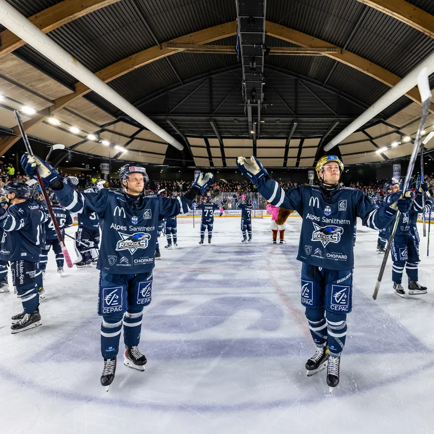 To ishockeyspillere i blå trøyer står på isen og hever stavene sine mens en heidundrende folkemengde er synlig i bakgrunnen.