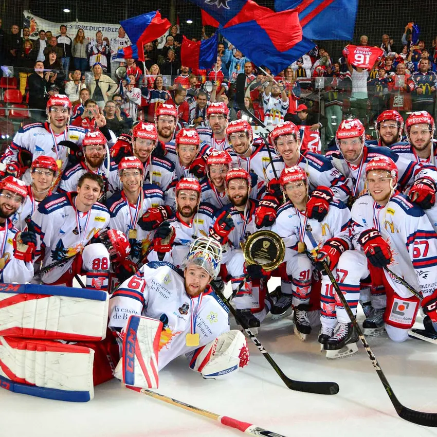 En gruppe ishockeyspillere i hvite trøyer med røde og blå detaljer poserer stolt med medaljer og et trofé på isen.