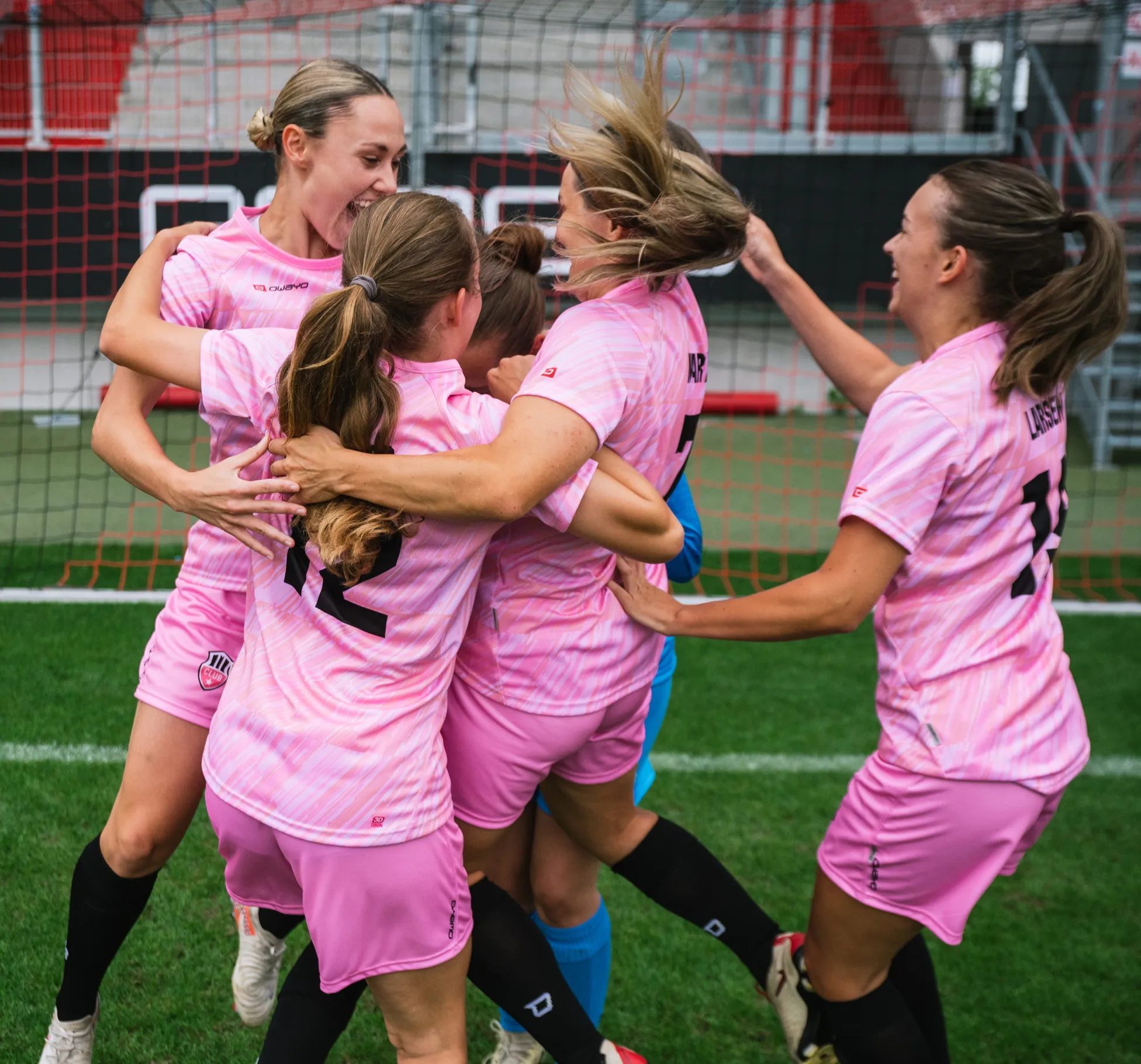 Fem kvinnelige spillere i rosa drakter feirer gledelig sammen på fotballbanen.