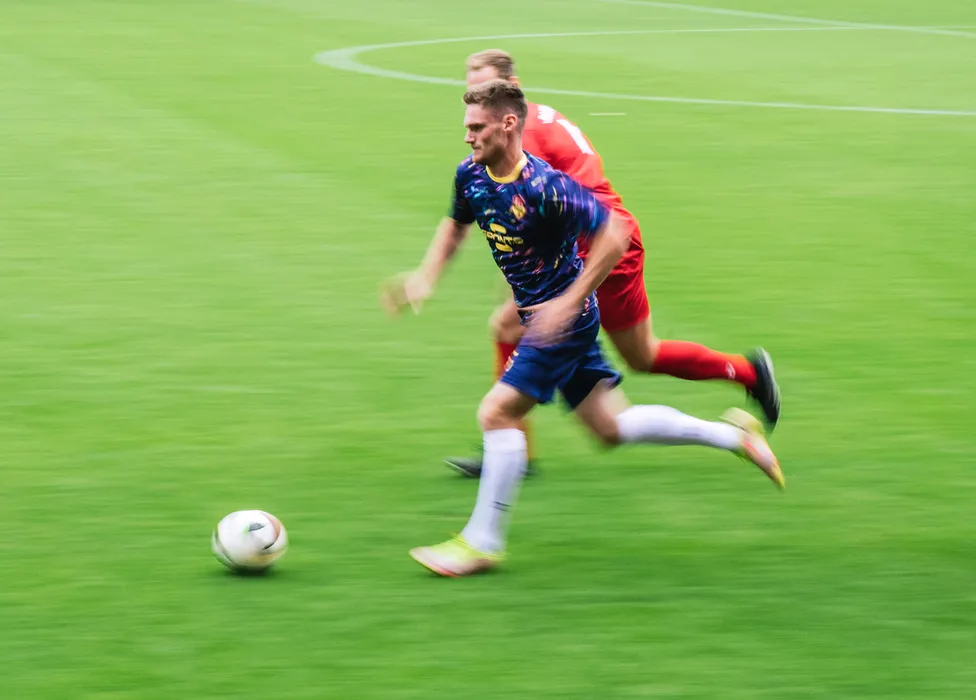 En fotballspiller i en blå drakt med fargerike mønstre løper raskt over den grønne banen mens en annen spiller følger tett etter.