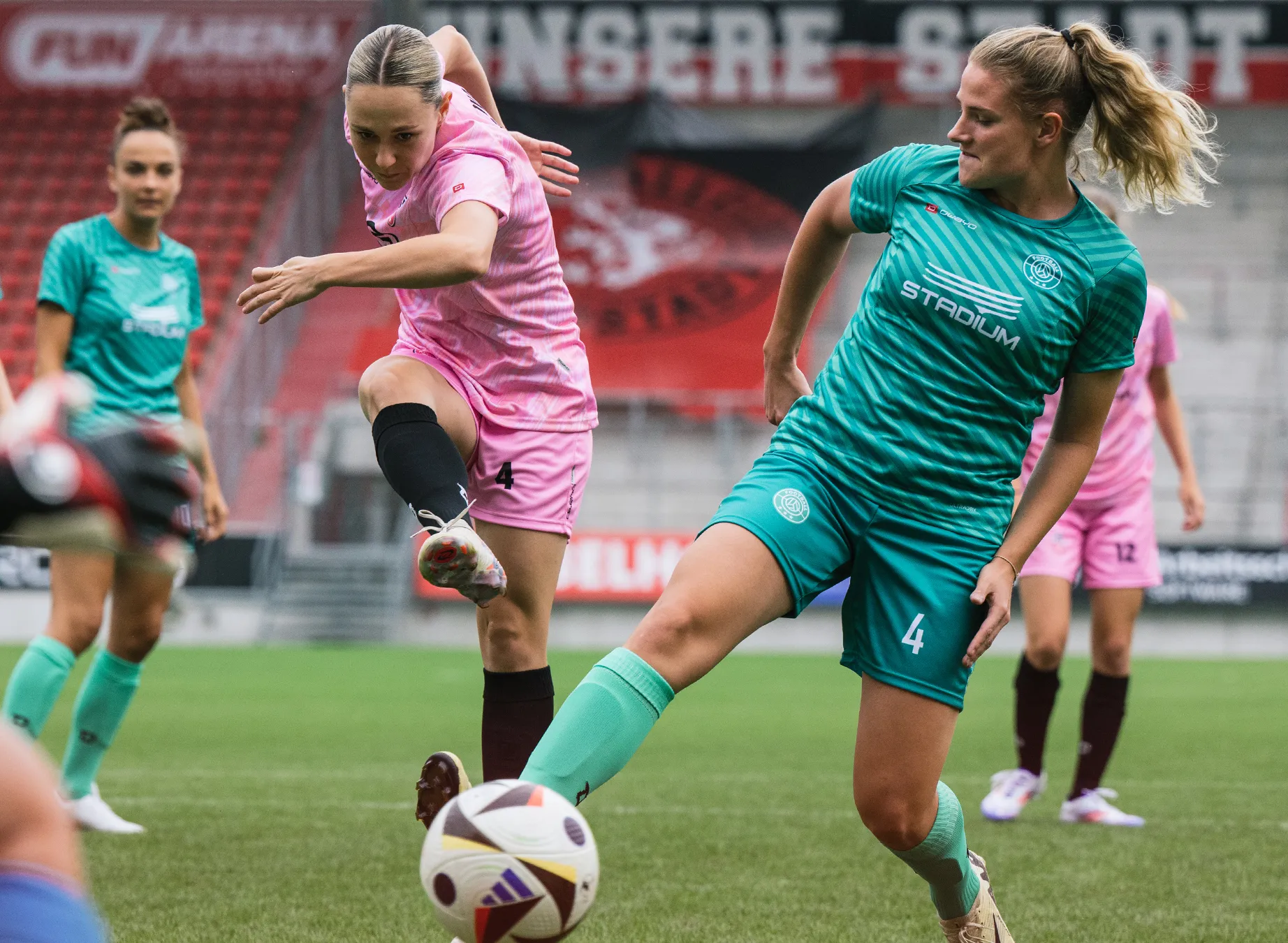 To kvinnelige fotballspillere i livlige trøyer, en i rosa og den andre i grønn, spiller på en gressbane.