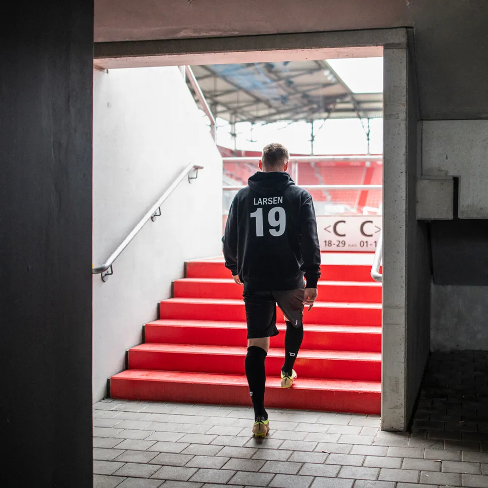 En atlet i hettegenser med nummer 19 på ryggen og svarte shorts går opp en trapp inn til et stadion.