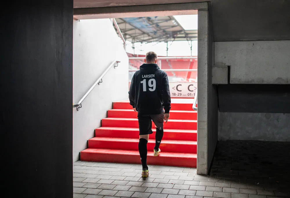 En fotballspiller i en svart hettegenser med nummer 19 forlater et stadionområde og går ned de røde trappene.
