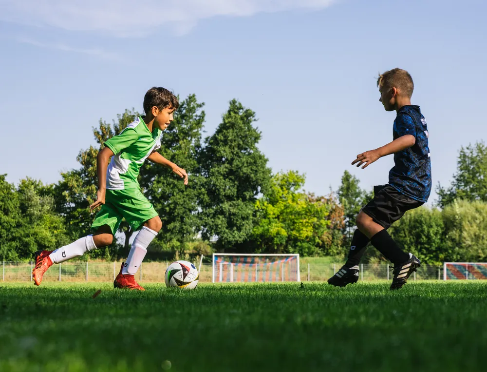 To barn spiller fotball på et grønt felt, den ene iført en grønn trøye og den andre i svart, mens de jager ballen.