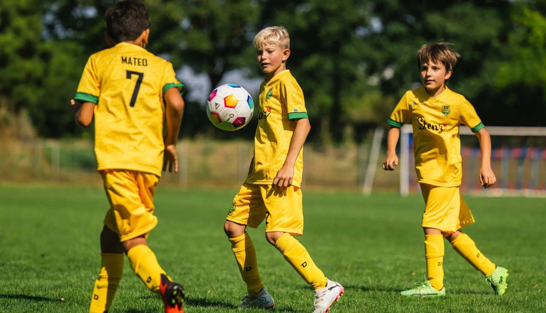 Tre barn i gule fotballdrakter spiller på en gressbane med en fargerik fotball.