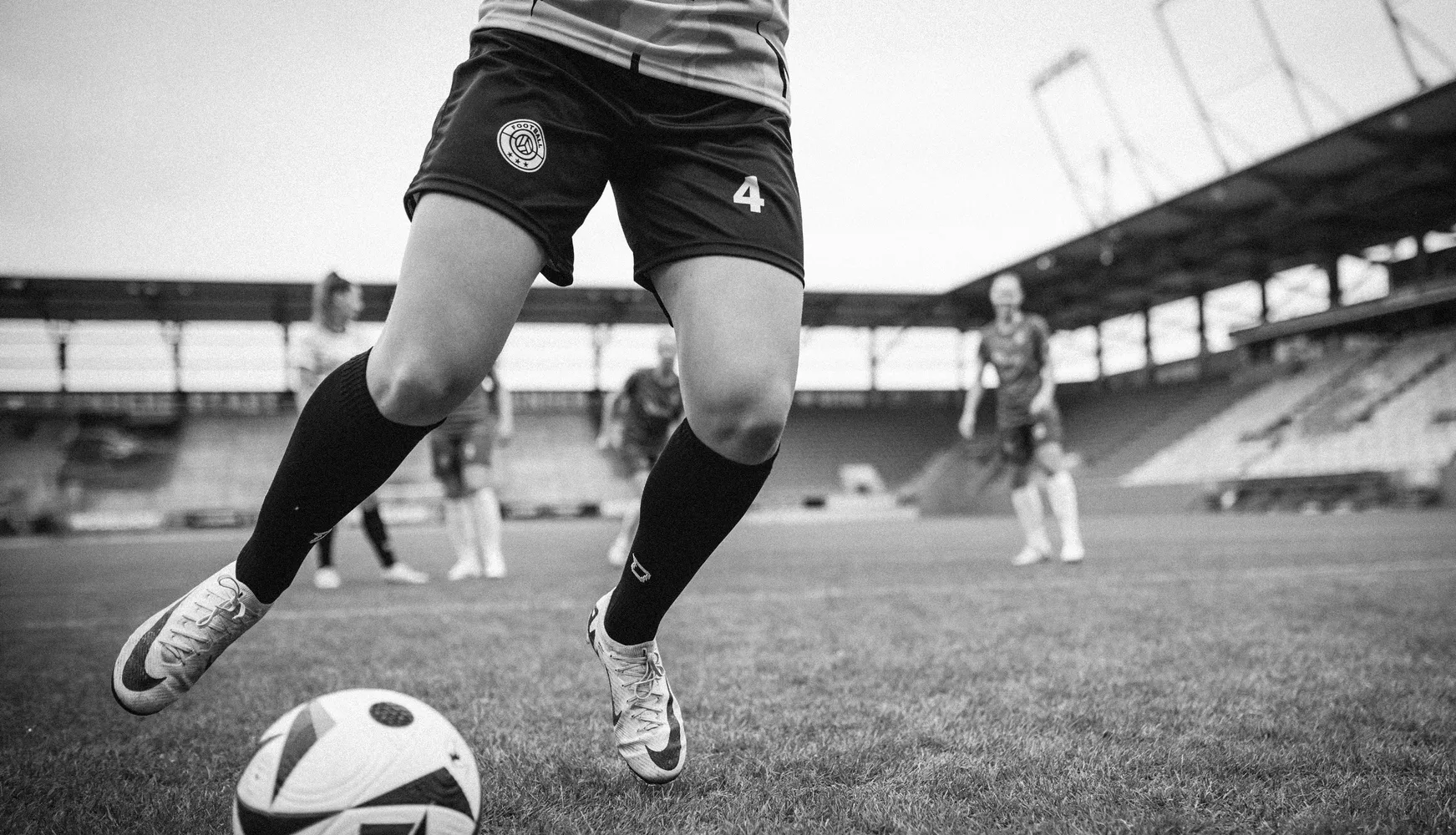 En kvinnelig spiller i korte fotballshorts og lange sokker sparker en ball på en fotballbane.