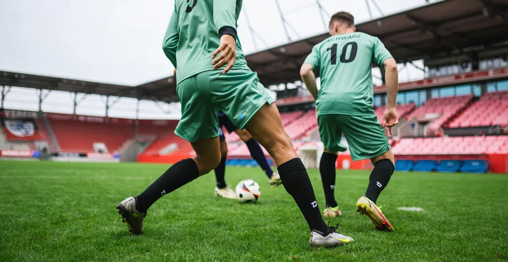 To fotballspillere i lysegrønne trøyer og svarte sokker som trener på en gressbane i en stadion.