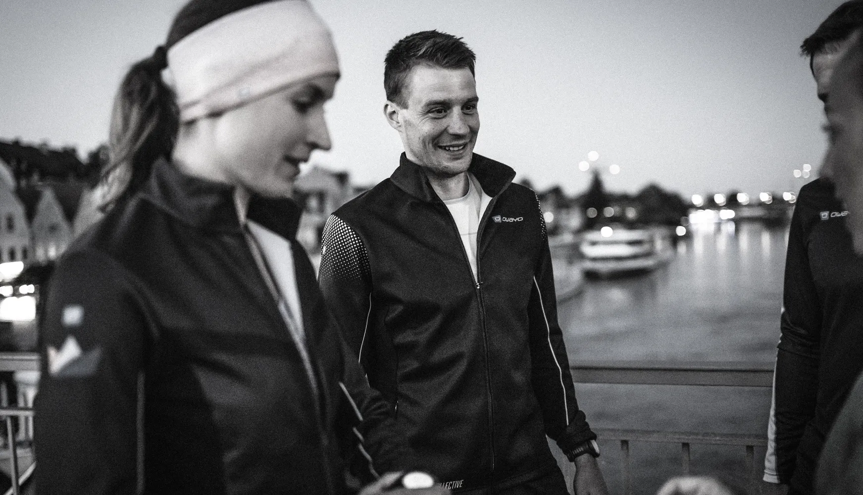 Tre personer i sportsklær står på en bro, mens de samtaler og ser på omgivelsene.