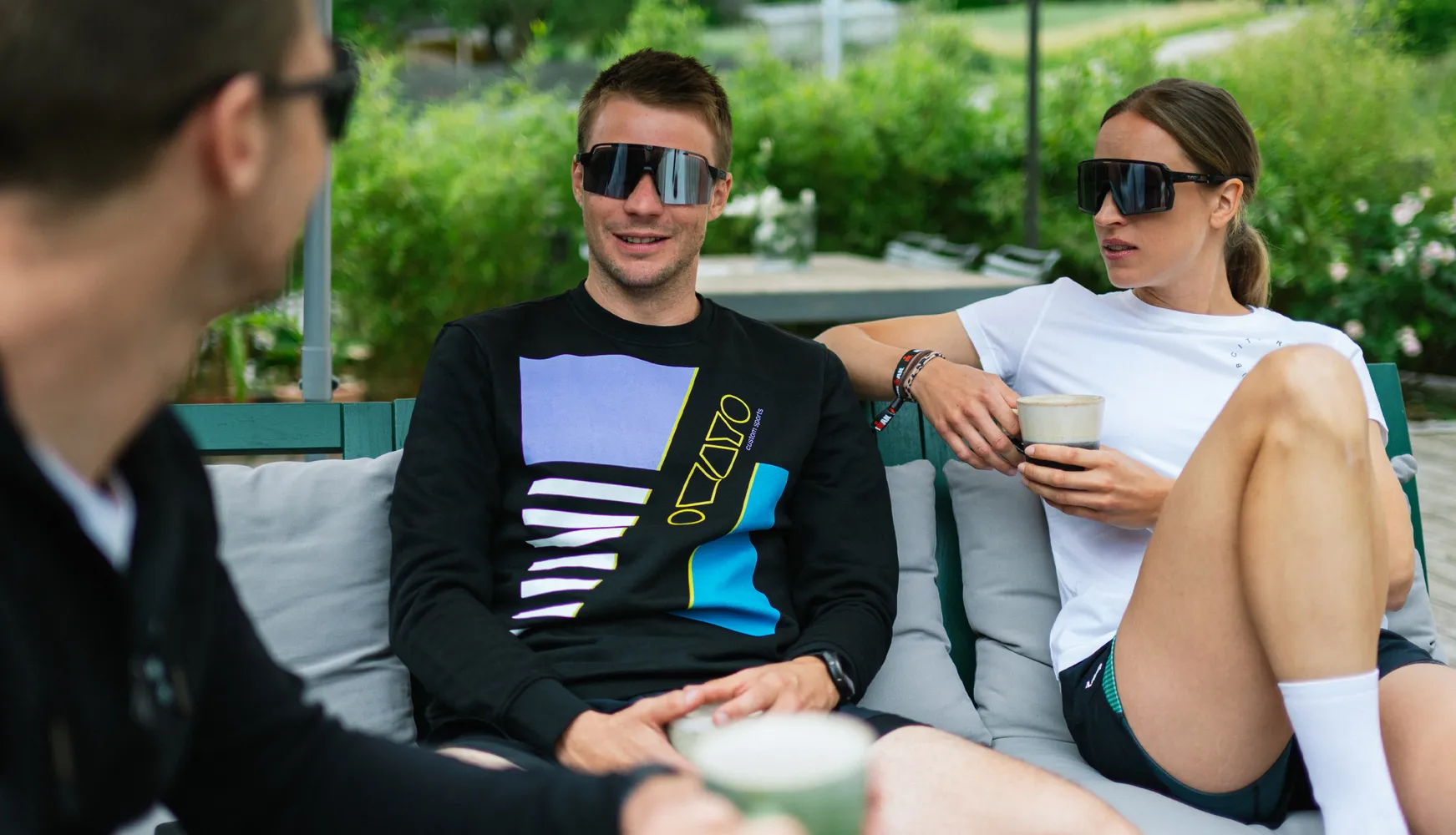 Tre personer i sporty antrekk slapper av på en ute-sofa mens de holder drikker.
