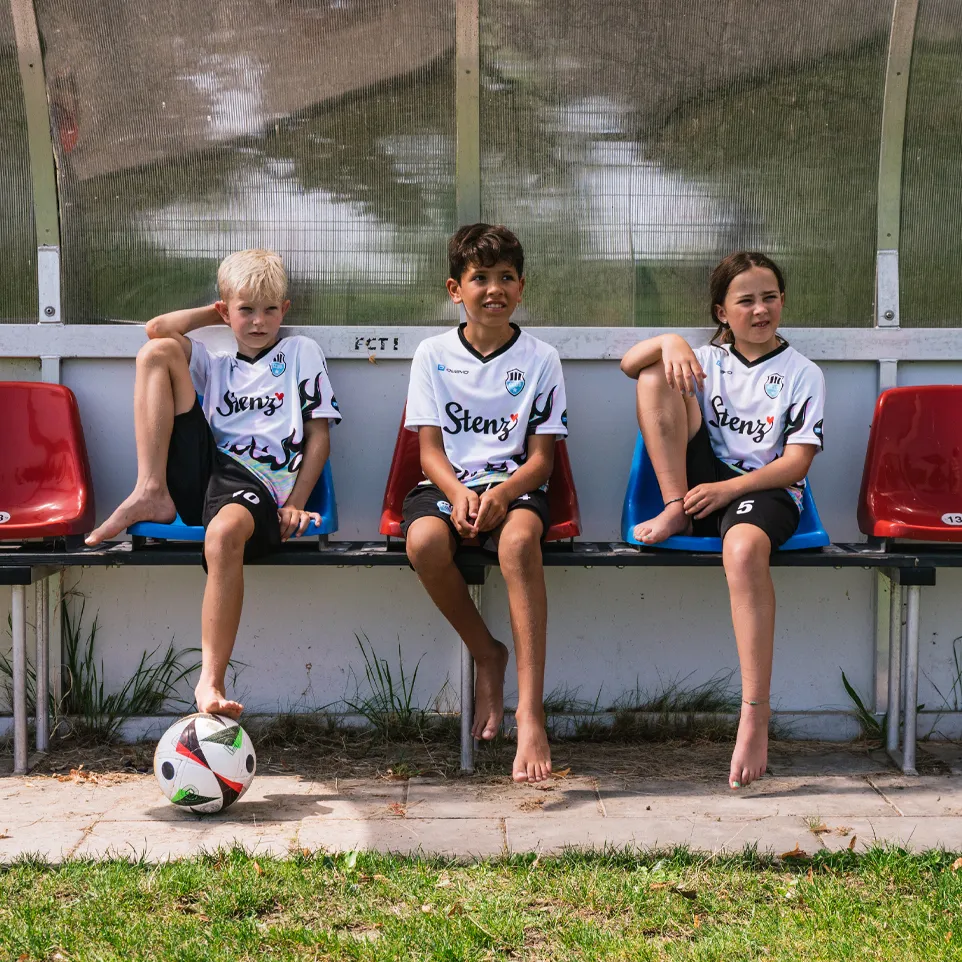 Tre barn i fotballdrakter og shorts sitter på en benk ved siden av banen, med en fotball foran dem.
