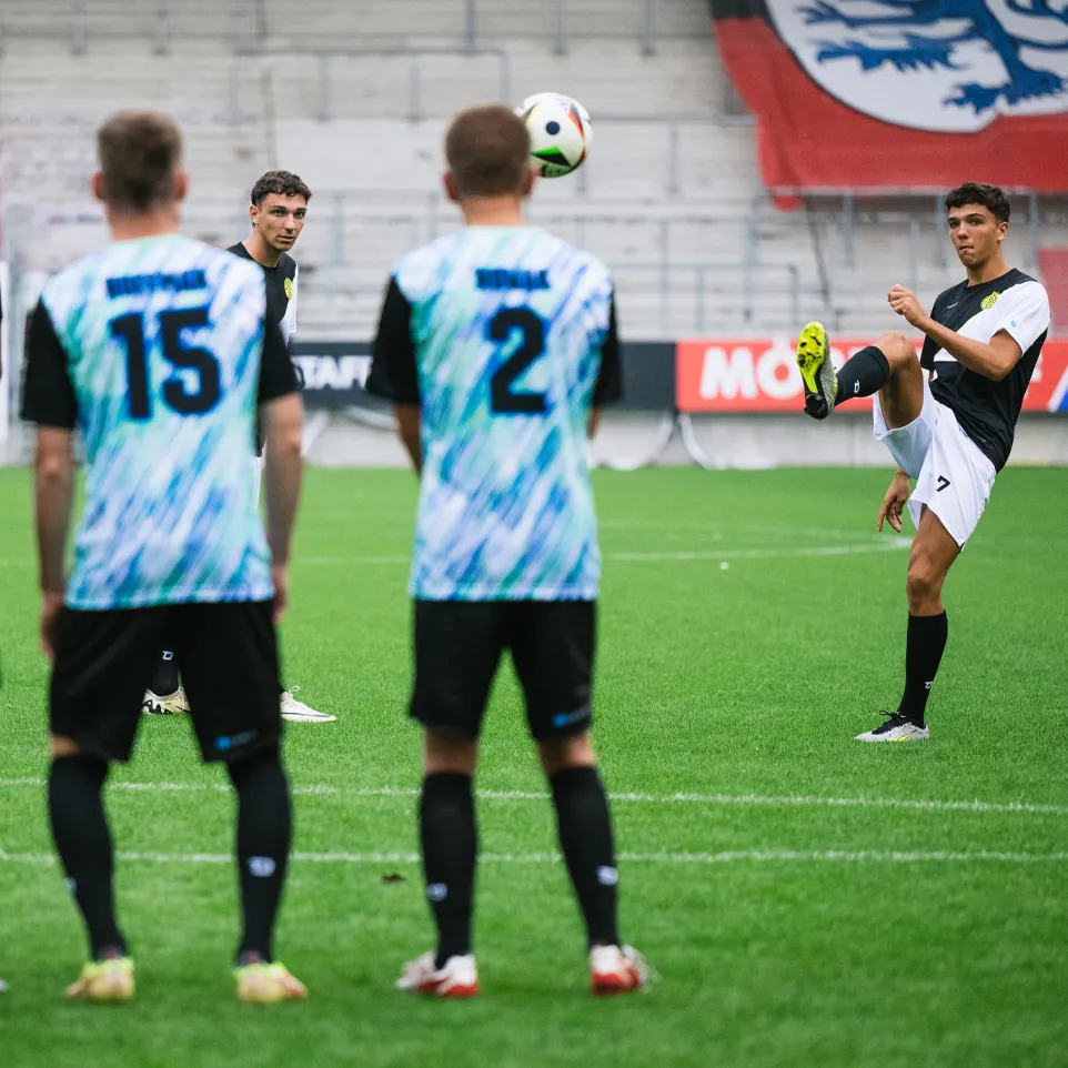 Tre fotballspillere i fargerike drakter står på banen mens en spiller sparker en pasning med høyre fot.