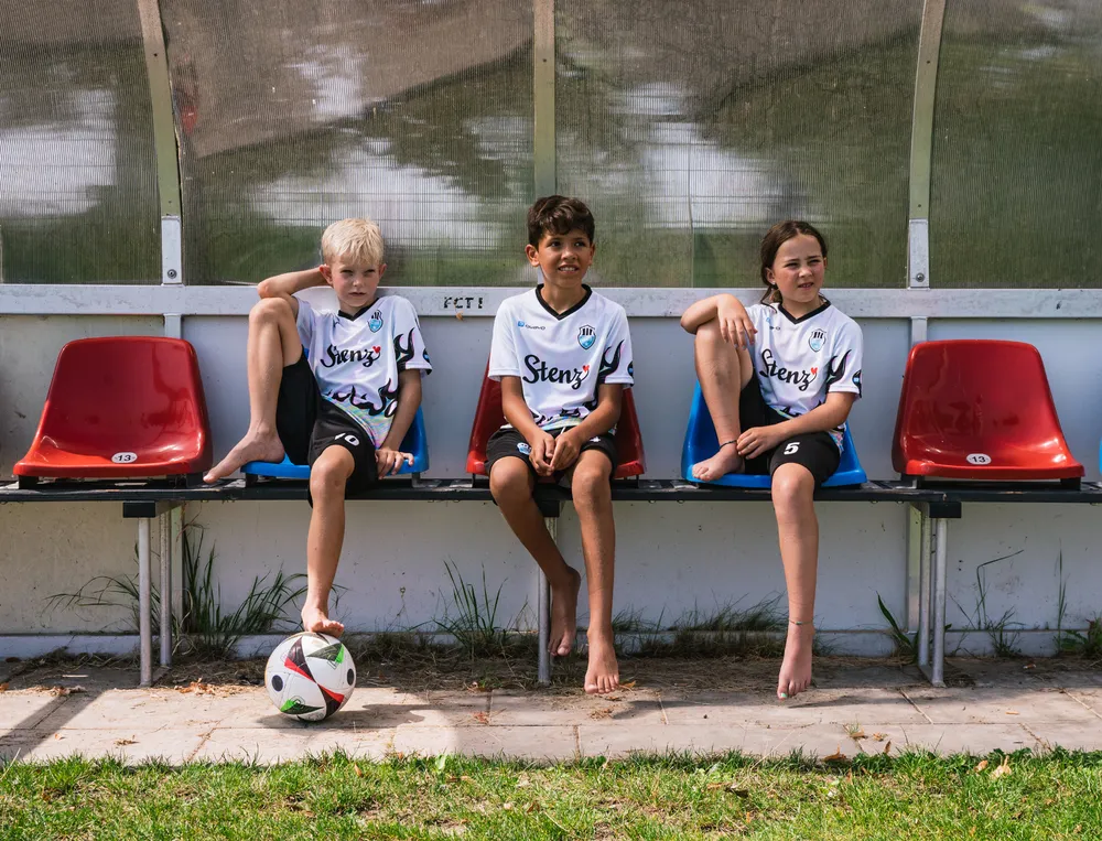 Tre barn sitter på en benk ved kanten av en fotballbane, iført tilpassede fotballdrakter med en fotball foran seg.
