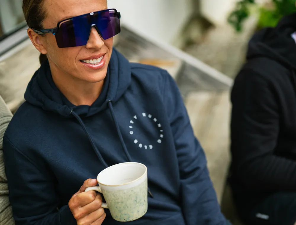 En person i en marineblå hettegenser med et logo holder en kopp kaffe og har på seg store solbriller.