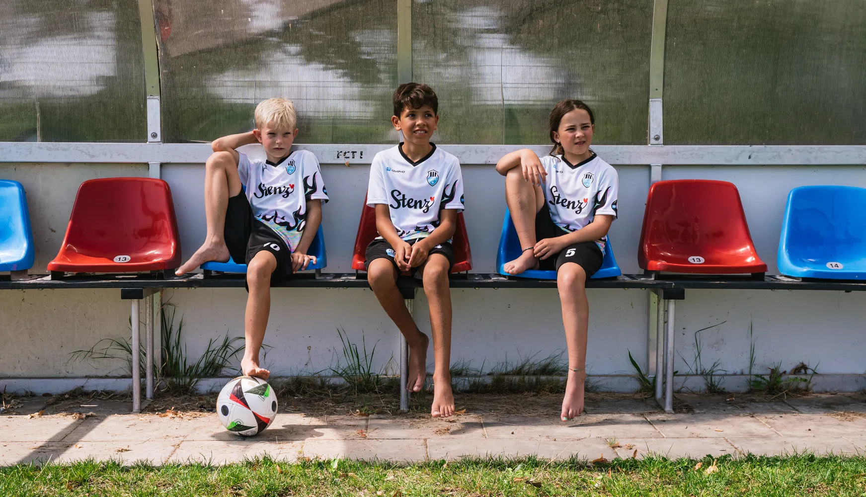 Tre barn i tilpassede fotballdrakter sitter på en benk med en fotball på bakken.