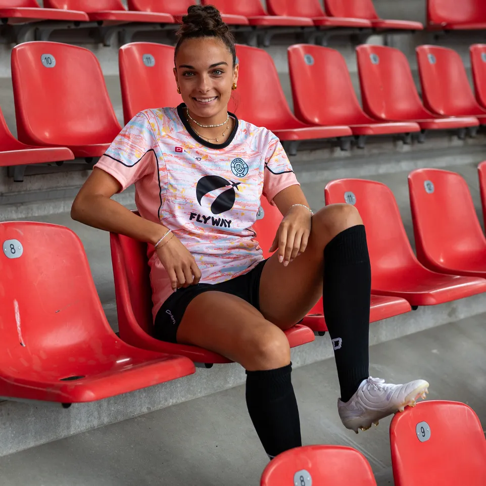 En smilende kvinne sitter på en rød stol i et stadion, iført en fargerik fotballdrakt og svarte sokker.