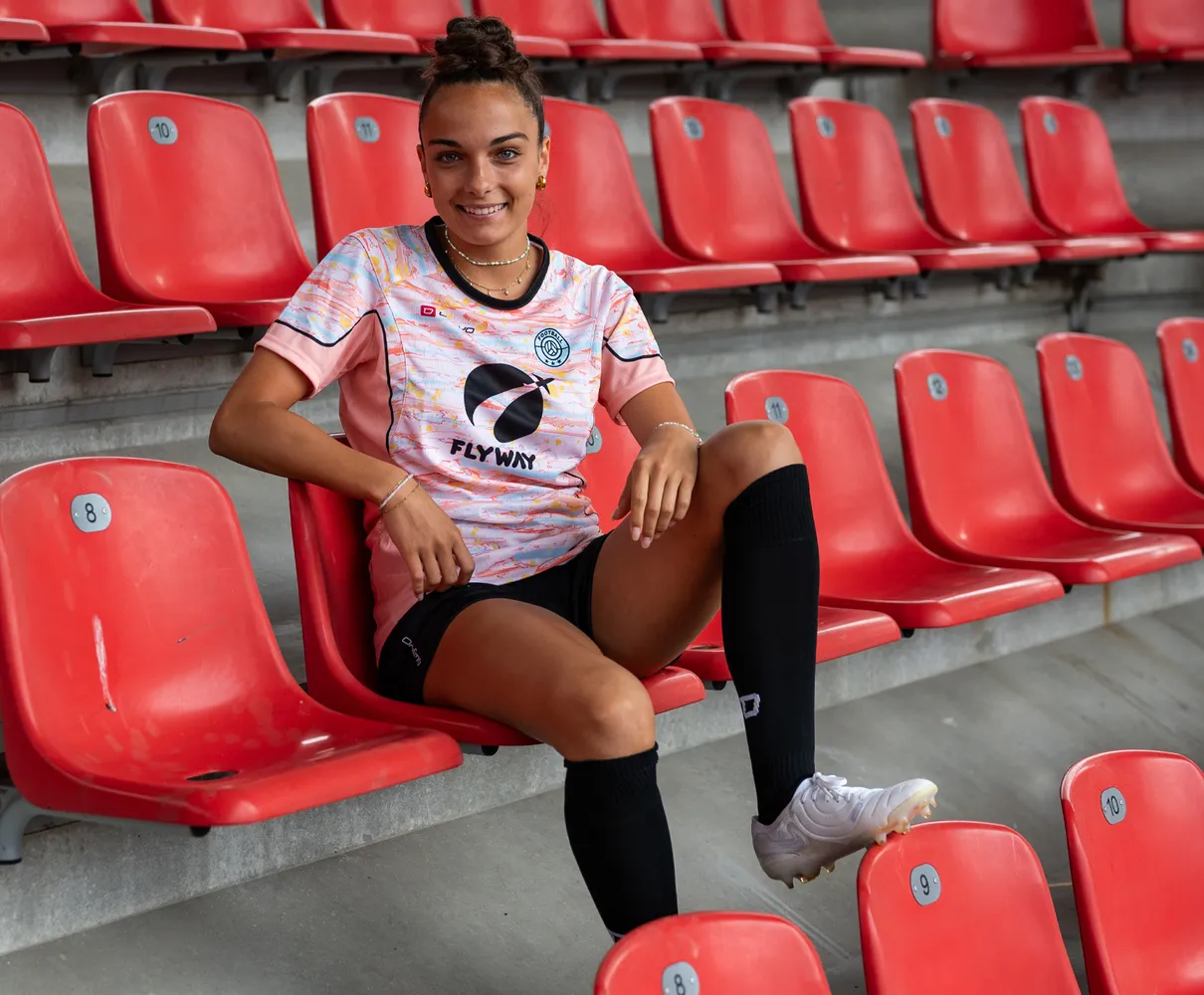 En ung kvinne sitter på en rød stadionstol iført en fargerik fotballdrakt og svarte sokker.