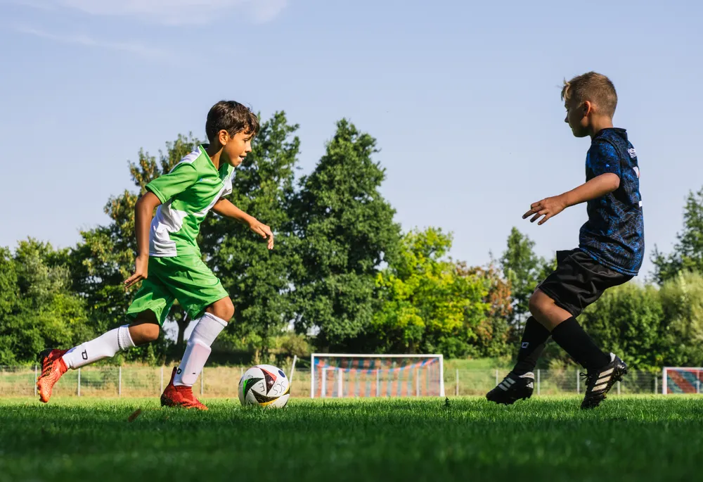To barn spiller fotball på en grønn bane, den ene i en grønn skjorte med hvite detaljer og den andre i en blå skjorte, mens de jager en ball.