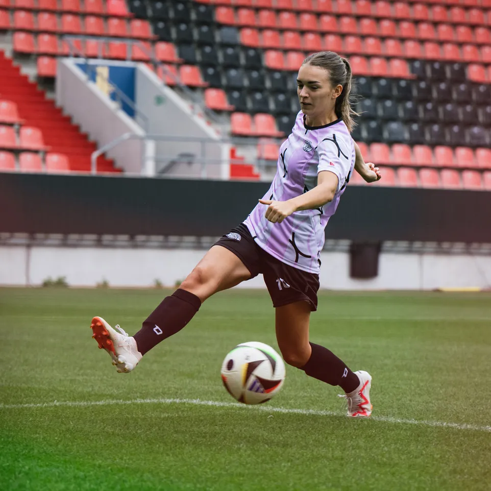 En kvinnelig spiller sparker en fotball på en stadionbane, iført en lilla trøye og svarte shorts.
