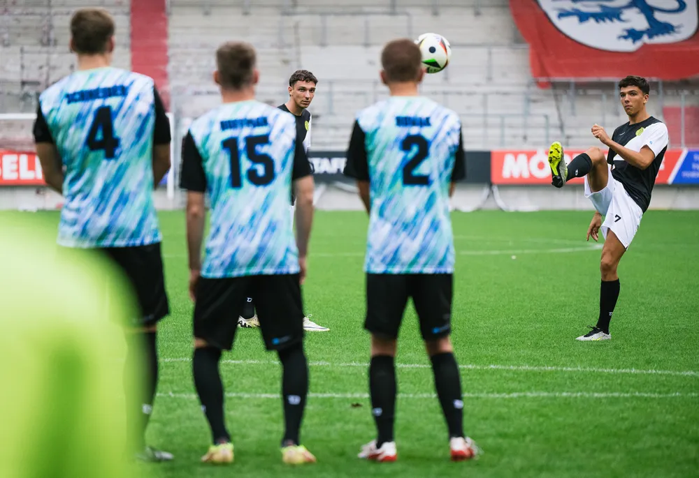En gruppe fotballspillere i fargerike trøyer står på banen mens en spiller løfter ballen med høyre fot.