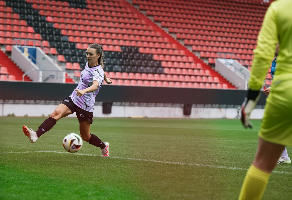 En kvinnelig fotballspiller i en lilla og svart drakt sparker en ball på en bane med tomme tribuner i bakgrunnen.