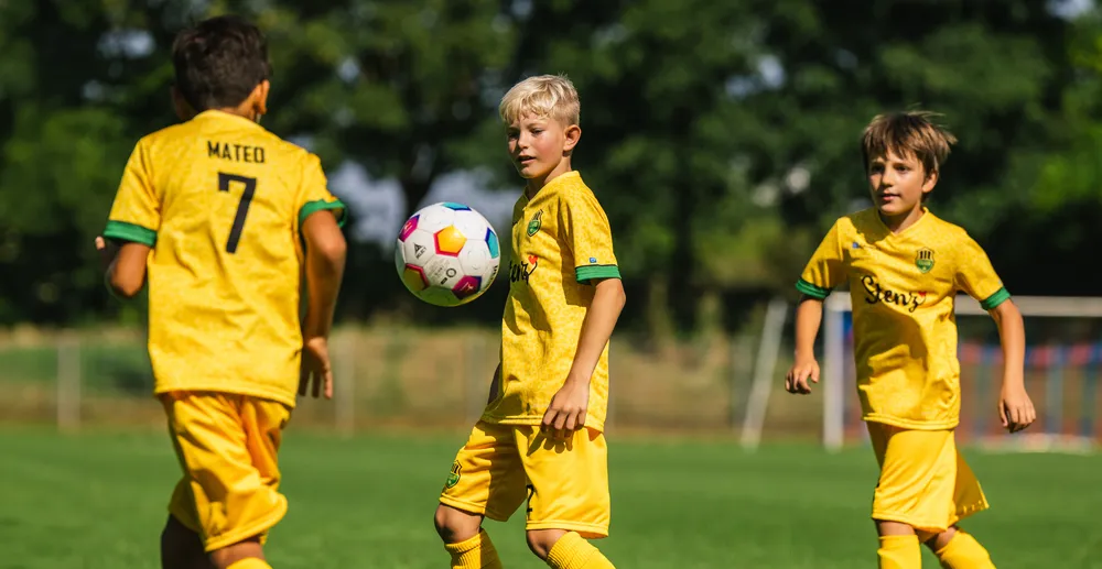 Tre barn i gule fotballdrakter spiller på en grønn bane, mens en av dem kontrollerer en fargerik fotball.