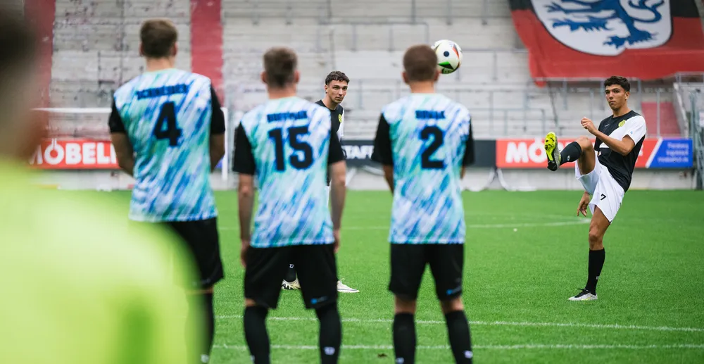 En gruppe fotballspillere i fargerike drakter står på banen mens en spiller løfter ballen med foten.