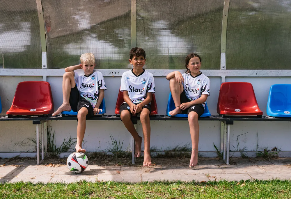 Tre barn i sporty fotballdrakter sitter på en benk ved siden av en fotball.