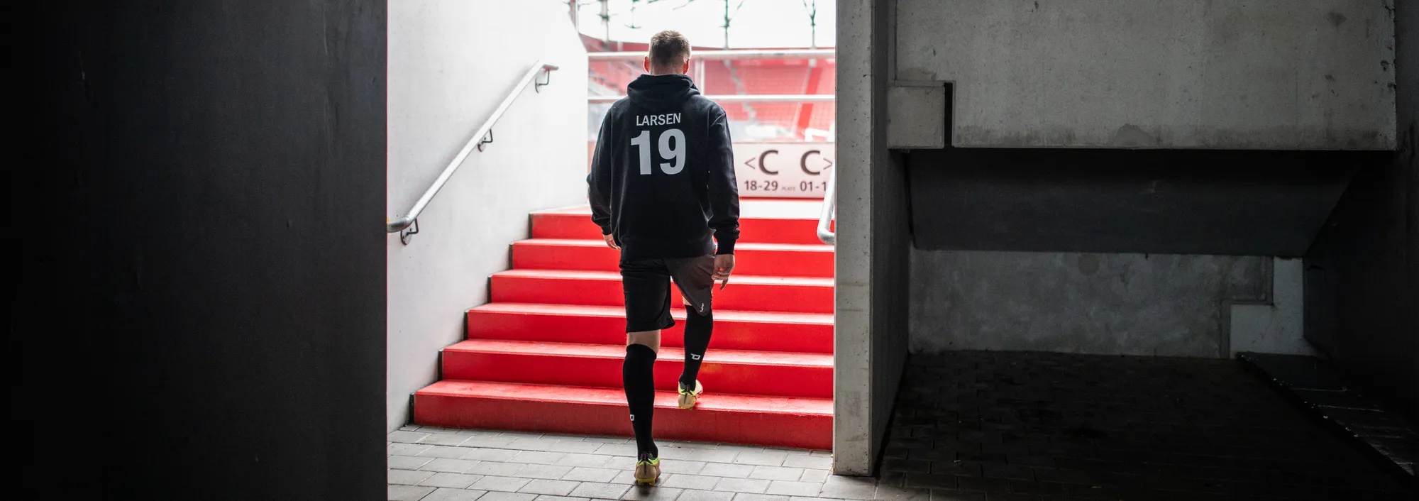 En spiller i en mørk hettegenser og shorts går opp de røde trappene fra en stadion.